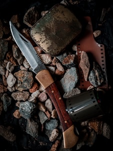A collection of objects including a hunting knife with a wooden handle, a weathered metal tin, a strip of photographic film, and a film canister are placed on a bed of rough pebbles.