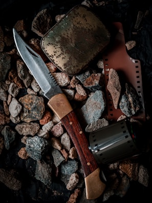 A collection of objects including a hunting knife with a wooden handle, a weathered metal tin, a strip of photographic film, and a film canister are placed on a bed of rough pebbles.