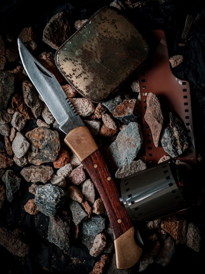 A collection of objects including a hunting knife with a wooden handle, a weathered metal tin, a strip of photographic film, and a film canister are placed on a bed of rough pebbles.