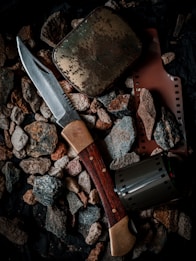 A collection of objects including a hunting knife with a wooden handle, a weathered metal tin, a strip of photographic film, and a film canister are placed on a bed of rough pebbles.