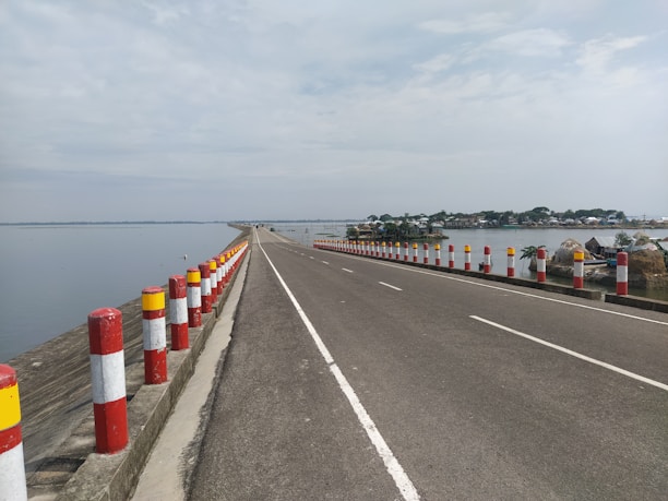 A long, straight road stretches out with water bodies on both sides. The road is flanked by red and white striped bollards. The sky is mostly clear with few clouds, and there are some houses and trees visible on the right side of the road in the distance. The road surface appears smooth and well-maintained.