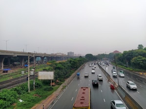 A highway lined with cars, including a large yellow bus, stretches into the distance. The road is bordered by greenery and an elevated metro or train line is visible on the left. The sky is overcast, suggesting a rainy or cloudy day, and the surface of the road appears wet.