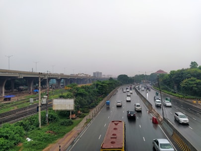 A highway lined with cars, including a large yellow bus, stretches into the distance. The road is bordered by greenery and an elevated metro or train line is visible on the left. The sky is overcast, suggesting a rainy or cloudy day, and the surface of the road appears wet.