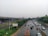 A highway lined with cars, including a large yellow bus, stretches into the distance. The road is bordered by greenery and an elevated metro or train line is visible on the left. The sky is overcast, suggesting a rainy or cloudy day, and the surface of the road appears wet.