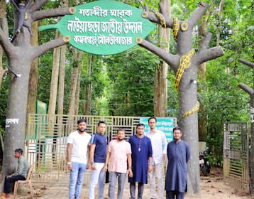 A group of six men are standing in front of a gated entrance to a national park with signage in Bengali. The entrance is flanked by artificial trees with a large model snake coiled around one. The area is surrounded by lush, green forest.