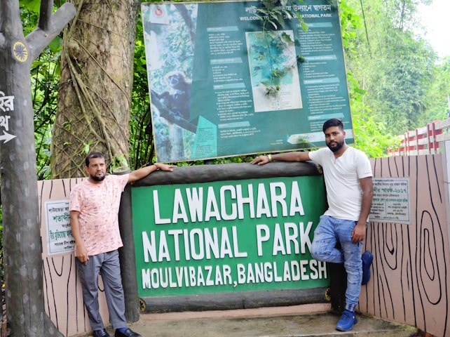 Two men standing next to the entrance sign of Lawachara National Park in Moulvibazar, Bangladesh. The area is lush with greenery and the sign is prominently displayed with informational boards above it.