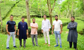 Six men are standing in a lush green tea garden, surrounded by tall trees and rows of tea plants. They are dressed in casual clothes, some wearing traditional attire. The atmosphere appears relaxed and serene, with vibrant greenery all around.