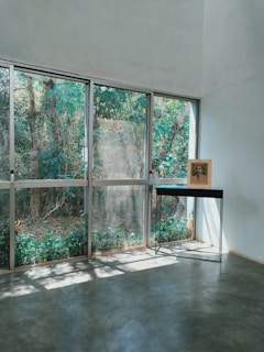 A minimalist interior space features large glass windows with a view of lush greenery outside. A single black table with slender metal legs stands near the windows, supporting a framed photograph or artwork. Sunlight streams through the windows, casting soft shadows on the smooth concrete floor.