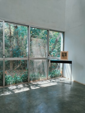 A minimalist interior space features large glass windows with a view of lush greenery outside. A single black table with slender metal legs stands near the windows, supporting a framed photograph or artwork. Sunlight streams through the windows, casting soft shadows on the smooth concrete floor.