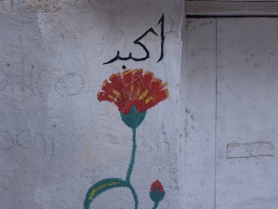 A simple wall mural featuring a red flower with yellow accents and a green stem, accompanied by Arabic script in black. The background is an uneven, light-colored surface that appears weathered.