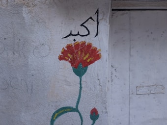 A simple wall mural featuring a red flower with yellow accents and a green stem, accompanied by Arabic script in black. The background is an uneven, light-colored surface that appears weathered.
