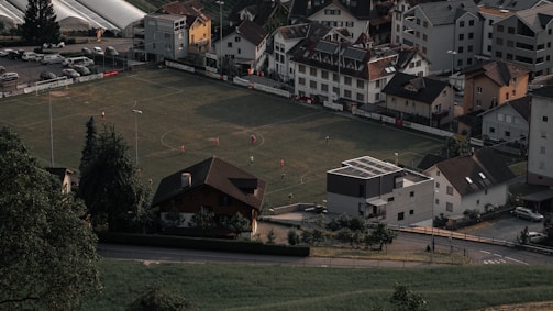 Aerial view of a small football field surrounded by residential buildings and a few cars parked nearby. Several players in red jerseys are visible on the field during a daytime match. The surrounding area includes greenery with trees and grass, as well as a quaint neighborhood setting with neatly arranged houses.