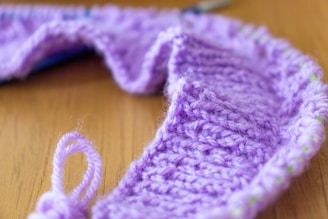 A close-up of a knitting project made with light purple yarn, showcasing a textured knit pattern. The piece is placed on a wooden surface, capturing the intricacies of the yarn fibers and the knit design.
