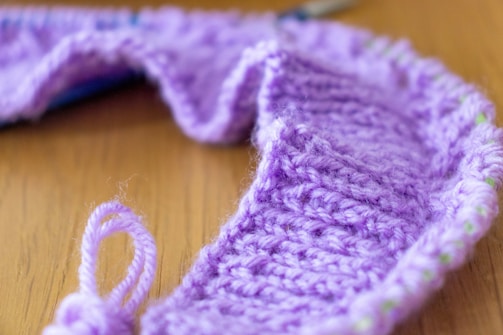 A close-up of a knitting project made with light purple yarn, showcasing a textured knit pattern. The piece is placed on a wooden surface, capturing the intricacies of the yarn fibers and the knit design.