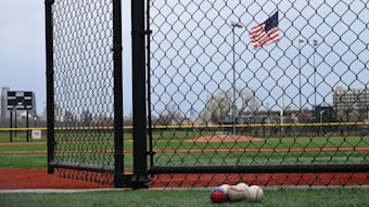 A chain-link fence in the foreground with a baseball field visible behind it. Three baseballs lie on the ground near the fence. An American flag is waving in the background, above the field. A scoreboard and light poles are also visible in the distance against a cloudy sky.