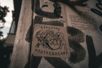 A weathered street pole covered in peeling stickers and graffiti. One prominent sticker features cartoon-like drawings of a cat, a mouse, and a dog holding a sphere, with text in Cyrillic script above and below the figures. The background includes partially visible instructions and cautionary text.