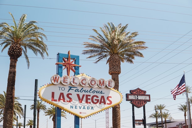 A vintage-style welcome sign displaying 'Welcome to Fabulous Las Vegas, Nevada' is surrounded by tall palm trees under a clear blue sky. An American flag waves on the right side, and a Harley-Davidson sign is visible in the background.
