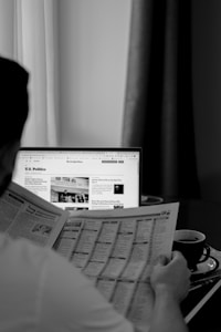 A person is reading a newspaper at a table with a laptop displaying a news website in the background. A cup is placed on the table, suggesting a relaxed reading moment. The scene is in black and white, enhancing the focus on the reading activity.