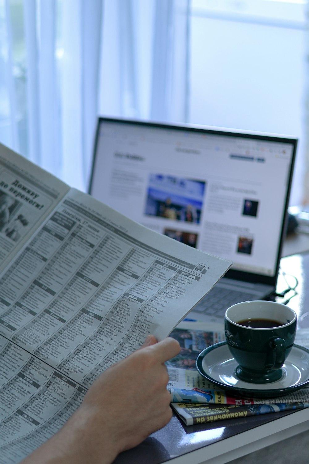 Una persona leyendo un periódico junto a una computadora portátil