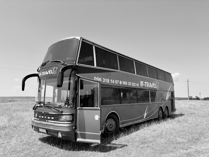 A black and white image of a large double-decker bus with the words 'IF-TRAVEL' displayed on the side along with a phone number. The bus is parked on a grassy field under a clear sky. The front entrance door of the bus is open, and there are six wheels visible on the side.