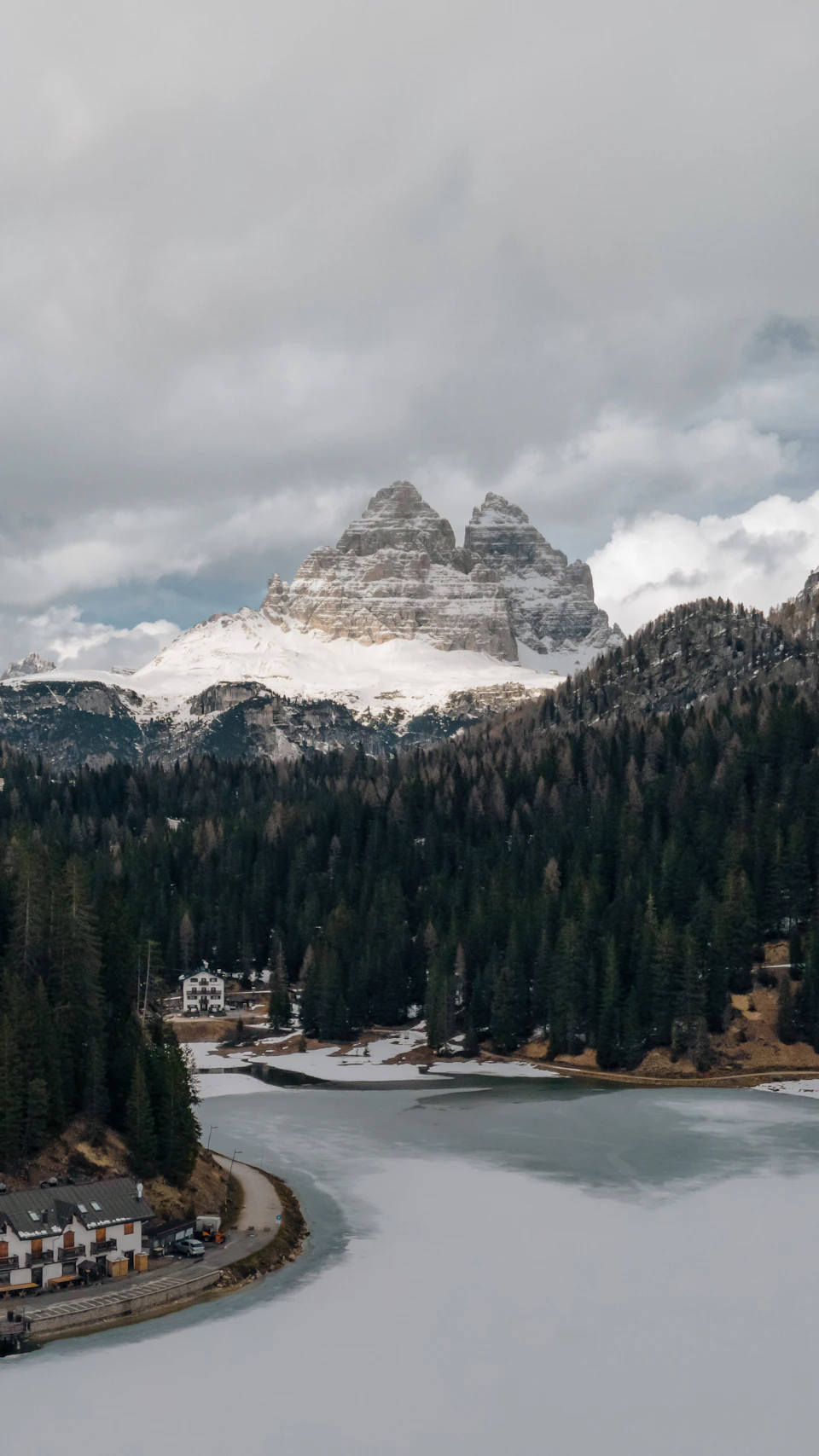 The image depicts a serene winter landscape with two snow-capped mountains standing tall against an overcast sky. The mountains are covered in a blanket of white snow, and their peaks are partially obscured by clouds. In the foreground, there is a small town nestled among the trees, with houses visible on both sides of a winding road that cuts through the scene.

The perspective of the image is from above, providing a bird's eye view of the mountains and the town below. The colors in the photo are predominantly gray and white due to the snow-covered landscape and overcast sky.