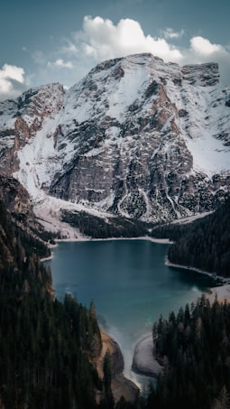 A stunning alpine landscape with a serene lake surrounded by dense forests, set against a backdrop of majestic snow-capped mountains under a partly cloudy sky.