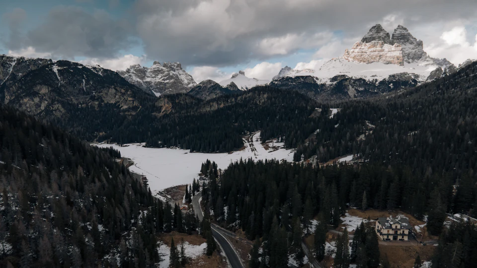 The image presents a breathtaking view of a mountainous landscape, with snow-capped peaks and a lake nestled amidst the trees. The sky above is filled with clouds, casting a soft light over the scene. In the foreground, there are two roads that lead towards the mountains, one of which is lined with trees on both sides. On the right side of the image, there's a small house surrounded by nature.