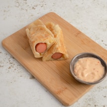 Two pieces of sliced pastry wrapped around sausages placed on a wooden cutting board, accompanied by a small bowl of creamy dipping sauce.