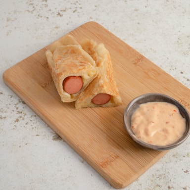 Two pieces of sliced pastry wrapped around sausages placed on a wooden cutting board, accompanied by a small bowl of creamy dipping sauce.