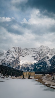 A grand hotel stands isolated by a frozen lake amidst towering snow-capped mountains. The cloudy sky adds a dramatic backdrop to the serene yet imposing natural landscape.