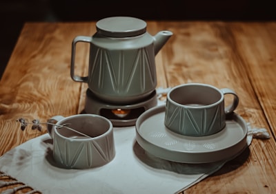 A ceramic tea set featuring a teapot and two cups is arranged on a wooden table. The teapot has a geometric pattern, and the cups are placed on a saucer with a similar design. A small decorative sprig lies near one of the cups, and a cloth is draped underneath.