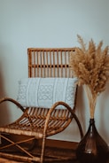 A wicker chair with a decorative pillow featuring geometric patterns is positioned next to a tall vase filled with pampas grass. The chair and vase are set against a plain wall, creating a cozy, minimalist atmosphere.