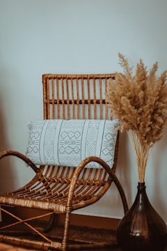 A wicker chair with a decorative pillow featuring geometric patterns is positioned next to a tall vase filled with pampas grass. The chair and vase are set against a plain wall, creating a cozy, minimalist atmosphere.
