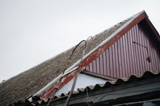 A close-up view of a sloped roof on a building, featuring corrugated material and a worn red wooden gable. Two metal bars with hooks are leaning against the roof. The sky is overcast, adding to the rustic and aged appearance of the scene.