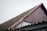 A close-up view of a sloped roof on a building, featuring corrugated material and a worn red wooden gable. Two metal bars with hooks are leaning against the roof. The sky is overcast, adding to the rustic and aged appearance of the scene.