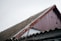 A close-up view of a sloped roof on a building, featuring corrugated material and a worn red wooden gable. Two metal bars with hooks are leaning against the roof. The sky is overcast, adding to the rustic and aged appearance of the scene.