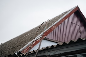 A close-up view of a sloped roof on a building, featuring corrugated material and a worn red wooden gable. Two metal bars with hooks are leaning against the roof. The sky is overcast, adding to the rustic and aged appearance of the scene.