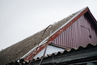 A close-up view of a sloped roof on a building, featuring corrugated material and a worn red wooden gable. Two metal bars with hooks are leaning against the roof. The sky is overcast, adding to the rustic and aged appearance of the scene.