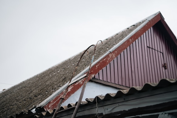 A close-up view of a sloped roof on a building, featuring corrugated material and a worn red wooden gable. Two metal bars with hooks are leaning against the roof. The sky is overcast, adding to the rustic and aged appearance of the scene.