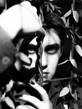 A black and white portrait of a person partially obscured by shadows and leafy vines. The intense gaze suggests a sense of mystery. The lighting creates high contrast, highlighting the texture of both the foliage and the person's features.