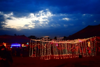 A nighttime community event illuminated by string lights, with people chatting and laughing.
