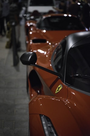 A line of sleek, high-performance sports cars with a focus on a red vehicle featuring an iconic prancing horse emblem. The image suggests a display or gathering, possibly at a car event. The setting is dimly lit, adding a sense of exclusivity and allure to the scene.