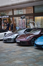Several luxury sports cars are parked in front of a showroom with large glass windows. There are people standing near the entrance, possibly security or staff, and a striking art installation is visible above one of the cars.