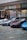 Several luxury sports cars are parked in front of a showroom with large glass windows. There are people standing near the entrance, possibly security or staff, and a striking art installation is visible above one of the cars.