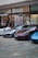 Several luxury sports cars are parked in front of a showroom with large glass windows. There are people standing near the entrance, possibly security or staff, and a striking art installation is visible above one of the cars.