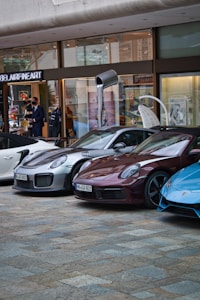 Several luxury sports cars are parked in front of a showroom with large glass windows. There are people standing near the entrance, possibly security or staff, and a striking art installation is visible above one of the cars.