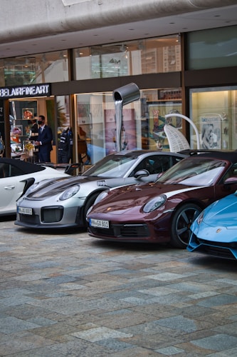 Several luxury sports cars are parked in front of a showroom with large glass windows. There are people standing near the entrance, possibly security or staff, and a striking art installation is visible above one of the cars.