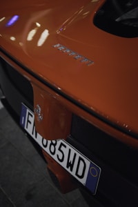 Close-up view of the back of an orange Ferrari car showcasing its branding and a European license plate. The Ferrari emblem and model name are prominently visible, with reflective light patterns on the vehicle’s glossy paint.