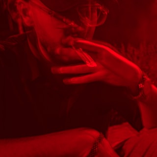 A person wearing glasses is smoking a cigarette while partially obscured in shadows. The lighting creates a red hue over the entire image, emphasizing the smoke and the relaxed posture of the individual.