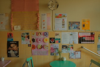 A wall filled with various posters and flyers in a cafe setting, featuring advertisements for events and products. The surface below has a small table with a potted plant and a green chair, creating a cozy and inviting atmosphere.
